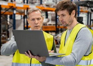 Two male workers wearing yellow safety vests are standing in a warehouse, closely reviewing data on a laptop. This image represents IT support services and manufacturing IT solutions being applied in a warehouse environment for inventory management and logistics.