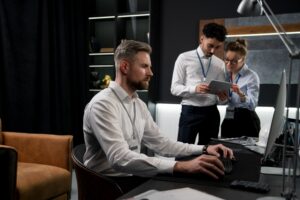 A man in a white shirt works intently at a computer in a modern office while two colleagues stand behind him, discussing information on a tablet. Represents IT services for law firms, focusing on managed IT services and cybersecurity solutions in a legal office environment.